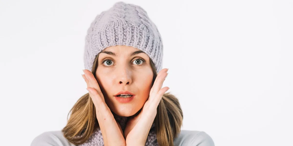 Woman in winter scarf and hat looking surprised, hand on face.